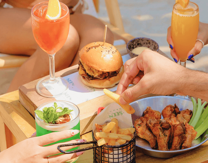 A spread at SEEN Eatery: a sliders board, chicken wings, fries, cocktails, and a juice on the table beside a pool daybed